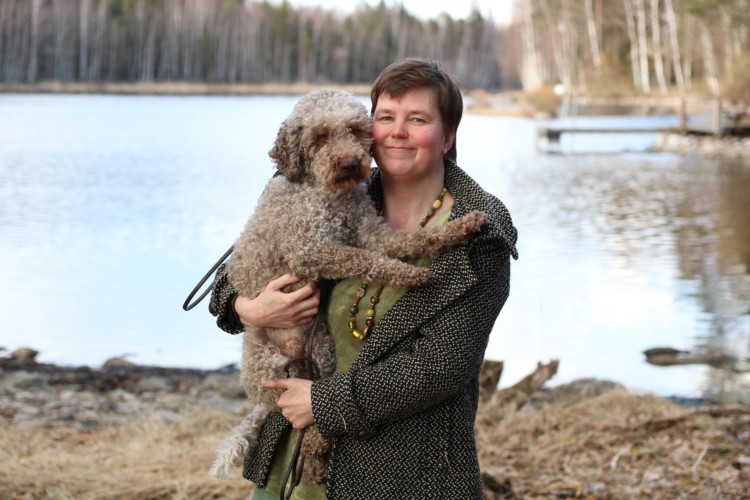 Reetta Tourunen seisoo ulkona lammen edessä koira sylissään.