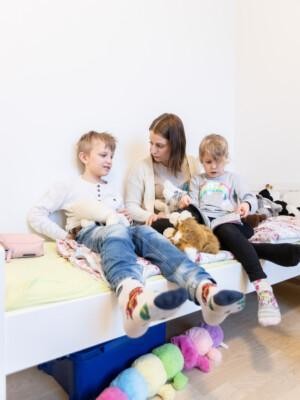Äiti ja kaksi lasta istuvat sängyllä. He lukevat kirjaa ja juttelevat. Sängyllä on lasten pehmoleluja.